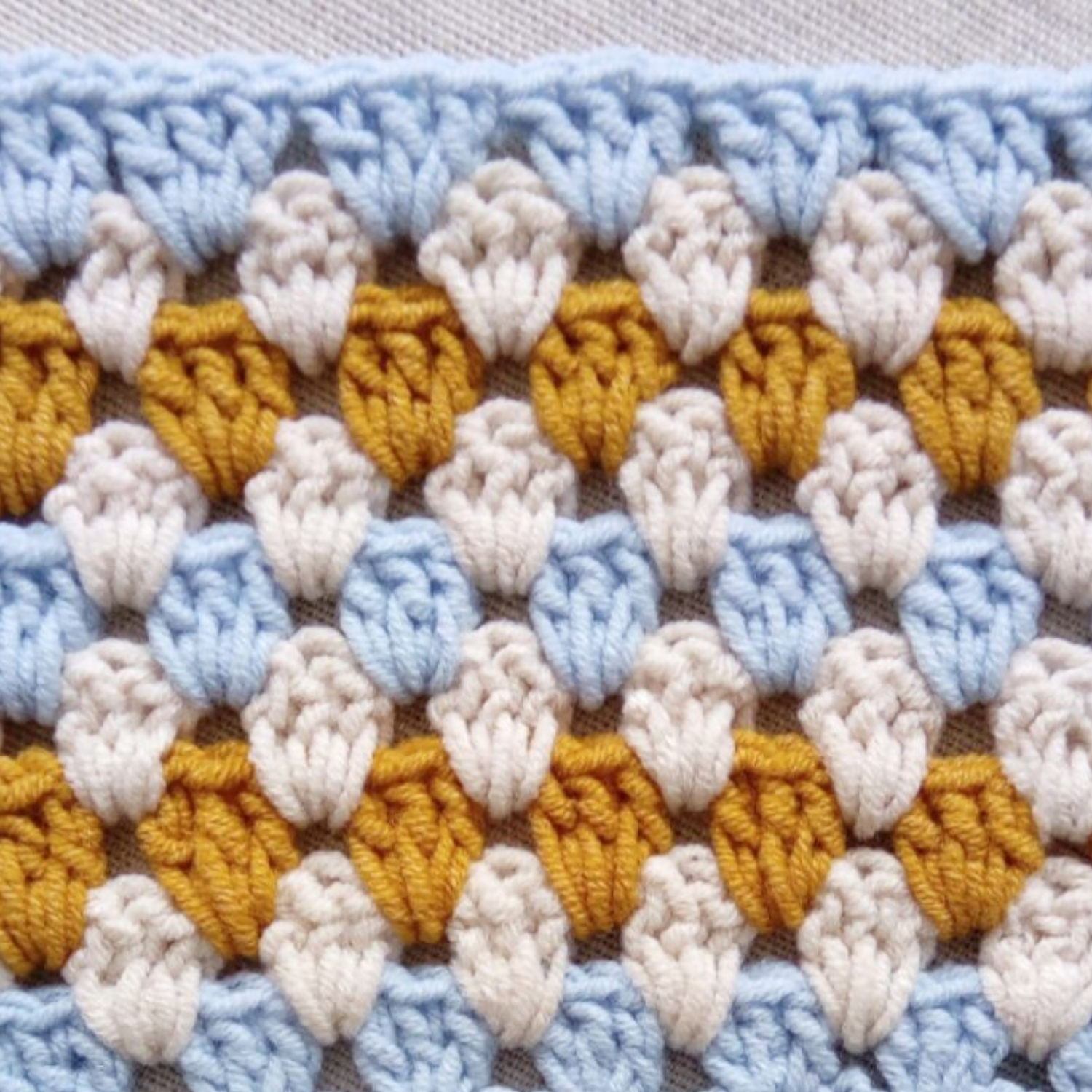 Crocheted pattern with rows of clusters in blue, white, and mustard yarn.