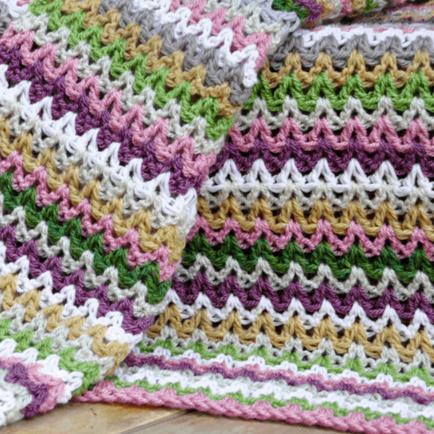 Close-up of a colorful crochet blanket featuring rows of multicolored zigzag patterns in shades of purple, green, white, and tan.