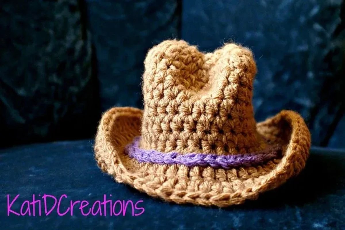 A crocheted cowboy hat designed for 0-3 months, featuring a purple band, rests on a dark blue surface. "KatiDCreations" is elegantly displayed in pink at the bottom left corner, showcasing delightful baby accessories.