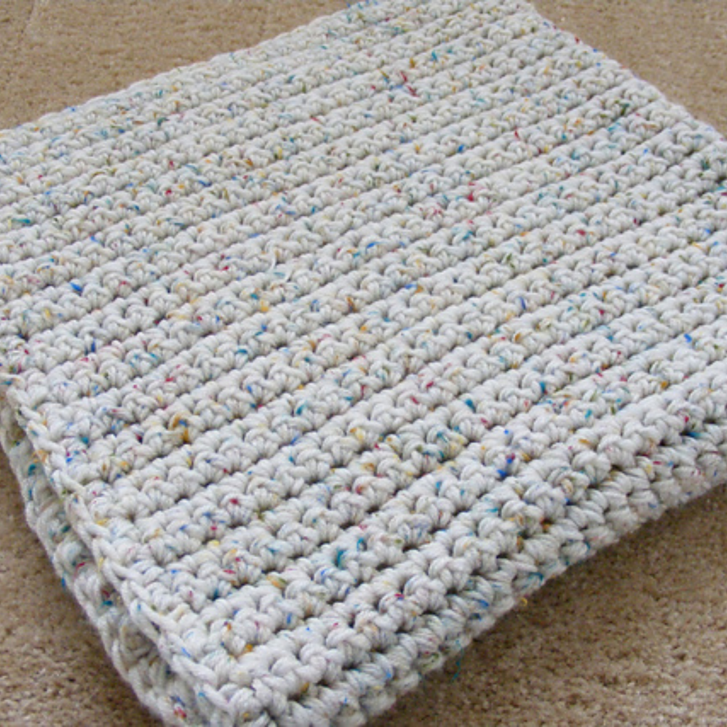 Folded crocheted blanket with a textured pattern and multicolored speckles is placed on a beige carpet.