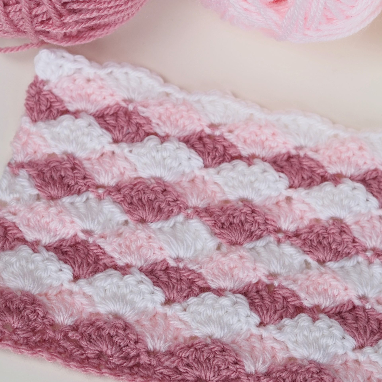 Crocheted fabric with a scalloped pattern in shades of pink and white, with skeins of yarn in similar colors nearby.