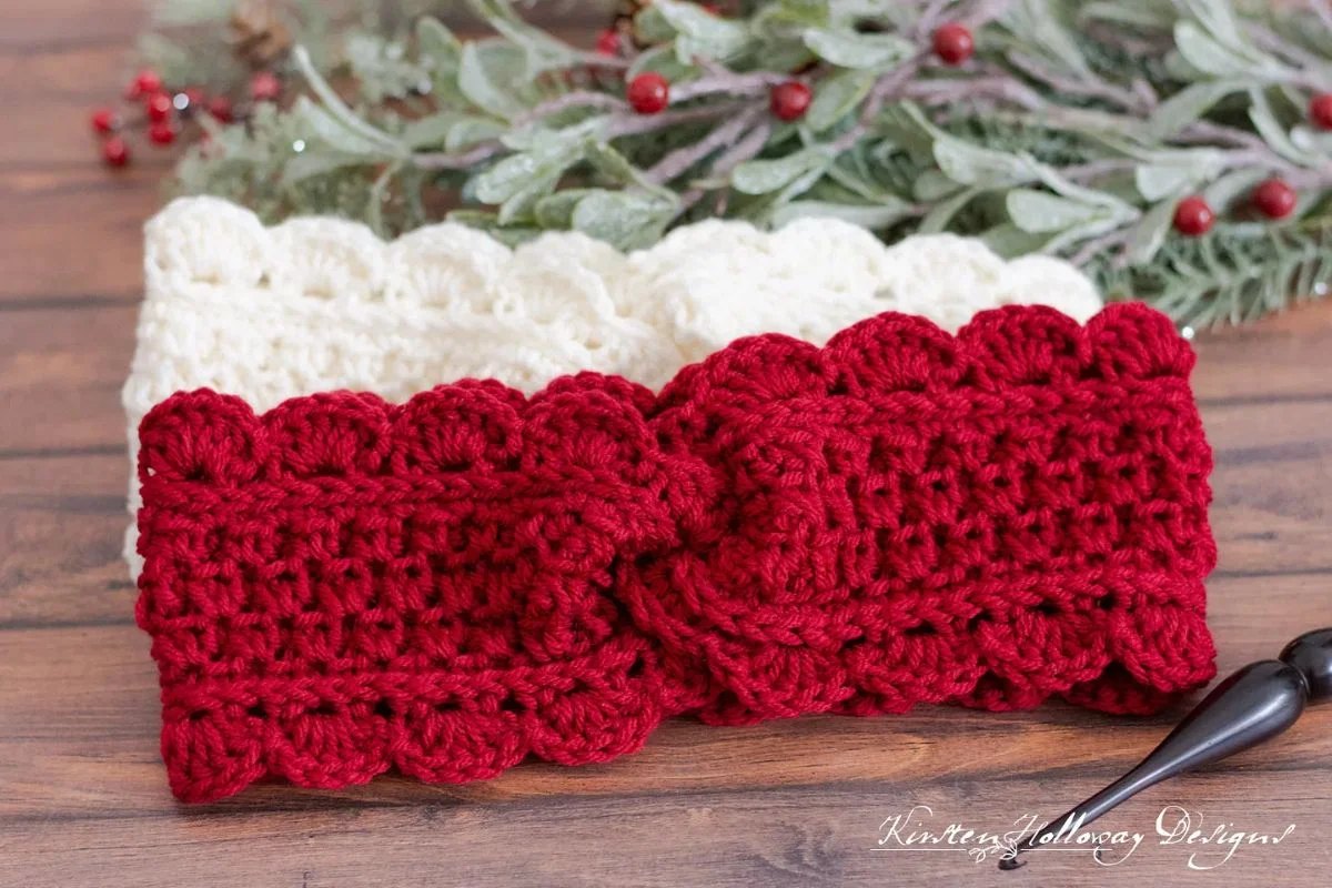 Two crocheted headbands, crafted with care, one in vibrant red and the other in soft white, are showcased against a rustic wooden surface. Lush greenery and red berries add a natural touch, reminiscent of the cozy warmth found in a handcrafted Tunisian Blanket.