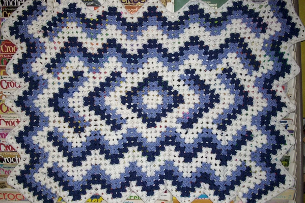 A crocheted blanket with a geometric pattern in blue, white, and purple hues is displayed against a backdrop of stacked magazines.