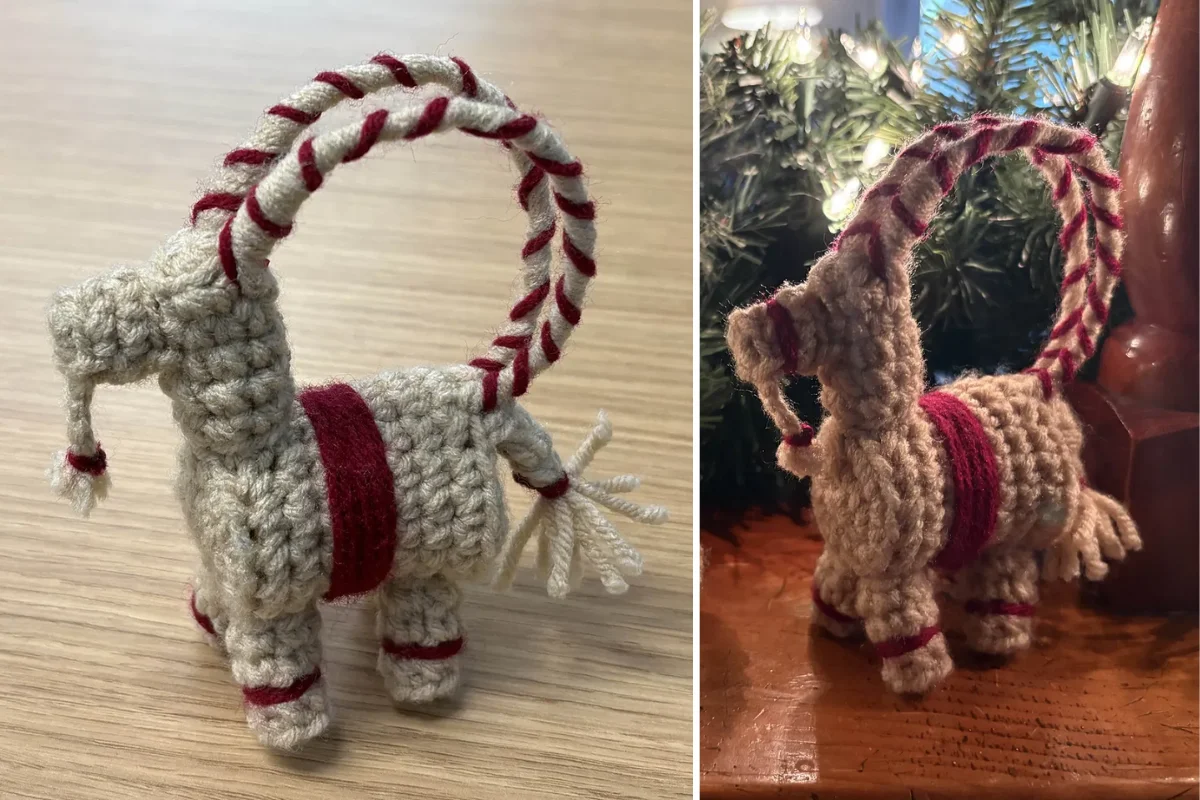 This charming crocheted Christmas Goat ornament, featuring red accents, rests against a wooden surface surrounded by festive greenery and twinkling lights.