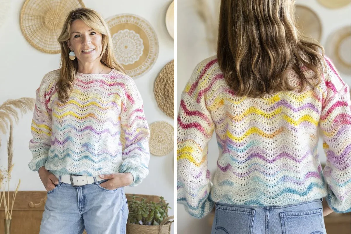 A person stands in front of a decorative wall adorned with woven baskets, wearing a multicolored crochet sweater with wavy patterns.