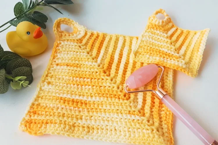 Two yellow striped knit dishcloths rest beside a pink facial roller and a rubber duck, all sitting cozily against the backdrop of a Hexagon Throw Cushion with lush greenery decorating the background.