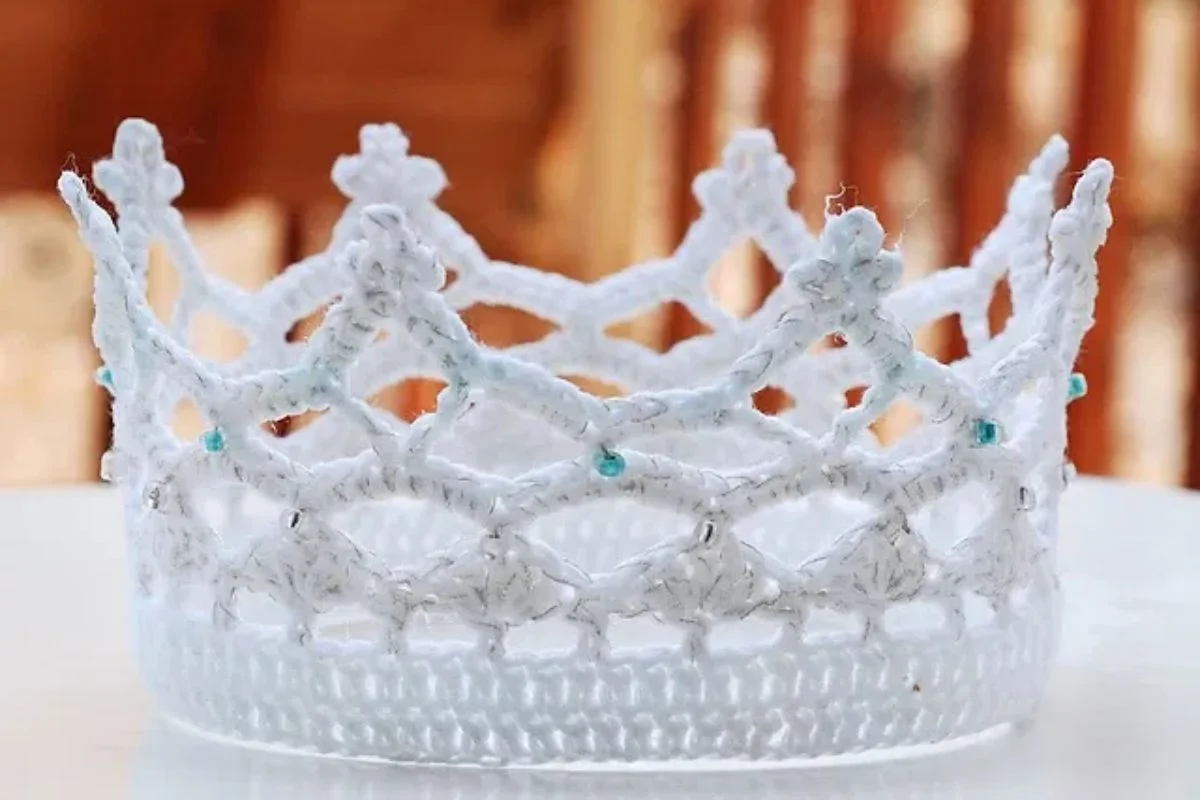 A handcrafted white crochet crown with intricate patterns and small blue beads.