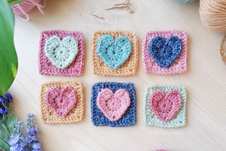 Six crochet heart squares in vibrant colors, arranged in two rows on a wooden surface, surrounded by lush green leaves and delicate purple flowers.