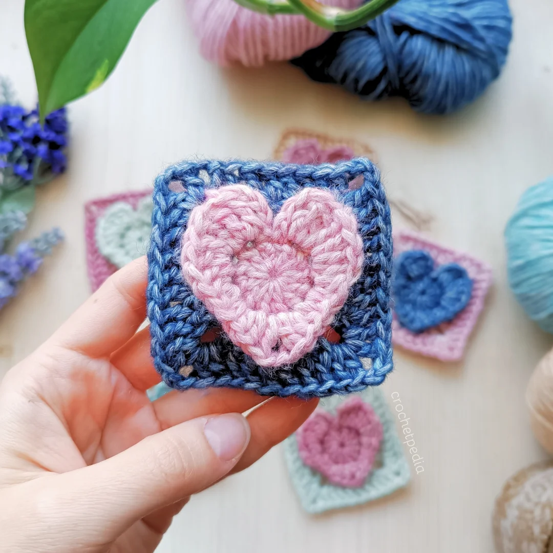 A hand holds a crochet heart square, featuring a pink heart at its center, set against a backdrop of yarn and other similar squares.
