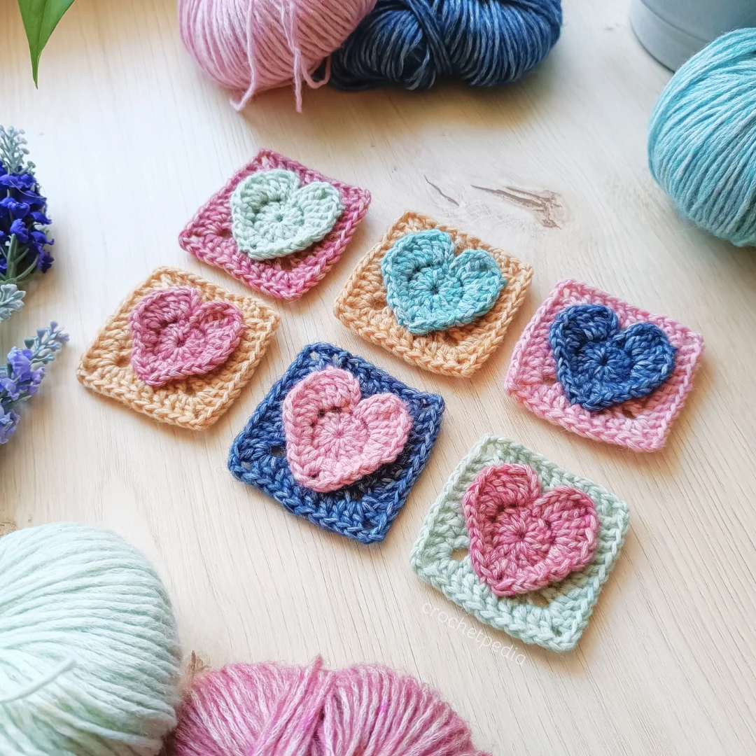 Crochet squares featuring delicate heart patterns in various colors are beautifully arranged on a wooden surface, surrounded by crochet heart-themed balls of yarn and small purple flowers.
