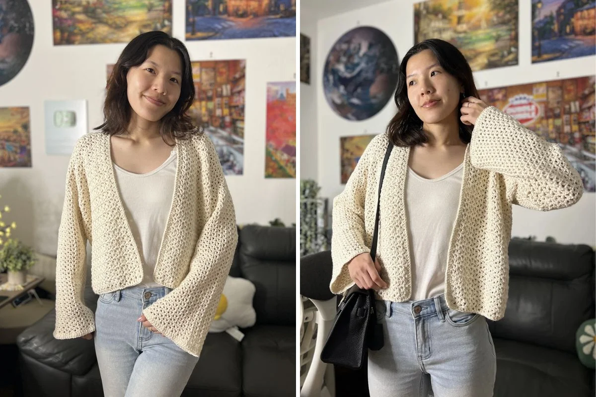 A person wearing a cream mesh crochet cardigan over a white shirt and jeans stands indoors in front of colorful paintings.