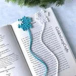 Two crocheted Snowflake Bookmarks, one blue and one white, are placed on an open book next to some pine branches.