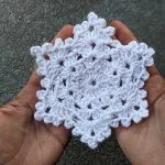 Two hands hold a delicate white crocheted snowflake ornament against a textured gray background.