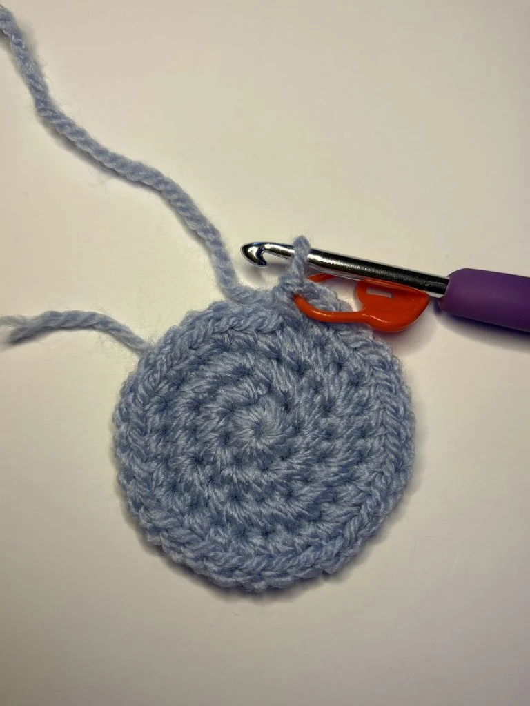 A crochet hook and stitch marker are attached to a circular piece of light blue crocheted yarn on a white surface, perfect for starting your next project like a crochet pumpkin pattern or even a whimsical crochet gnome.