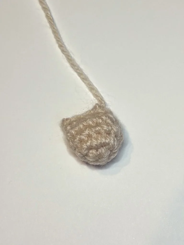 A small, beige crocheted piece with an attached yarn tail lies on a plain white surface, perfect as the base for a pumpkin crochet pattern or even a whimsical crochet gnome.