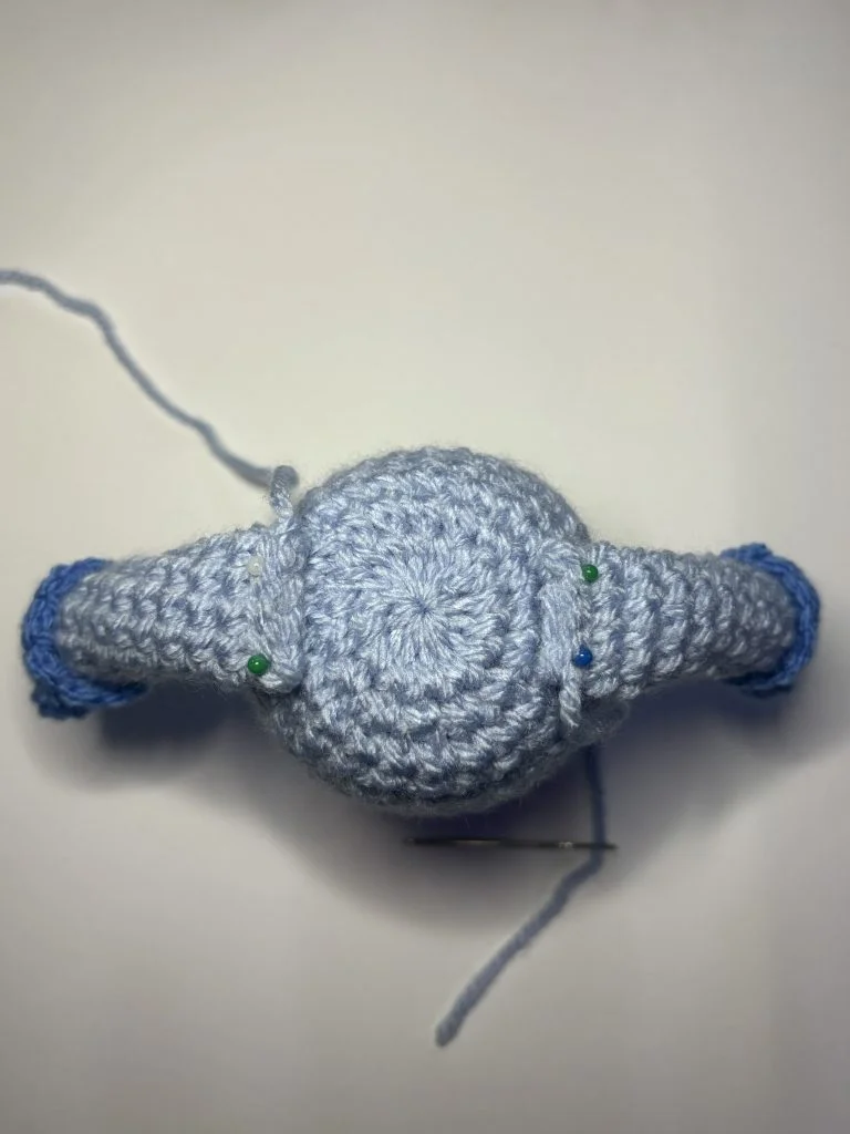 A top-down view of a crocheted blue object with two attached arms, held in place by green pins, and a needle with thread nearby—perfect for those following a crochet gnome or pumpkin crochet pattern.