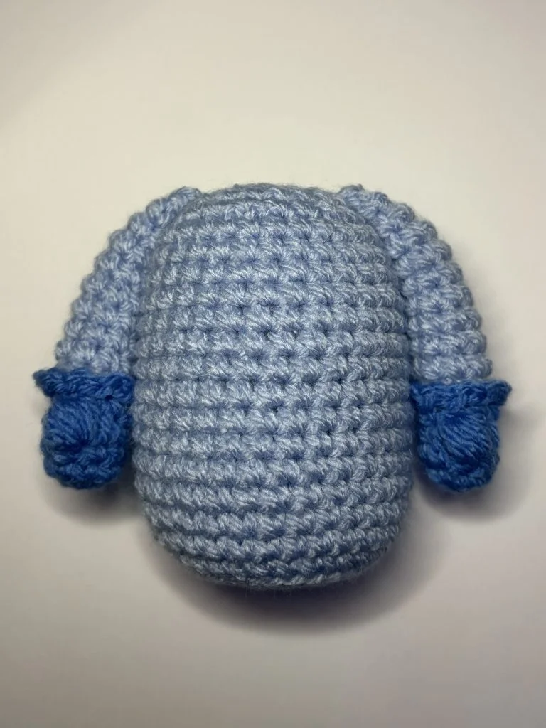 A crocheted blue oval shape with two short arms, each wearing a darker blue mitten, sits on a plain white background—perfect inspiration for your next free crochet pumpkin pattern or playful crochet gnome project.
