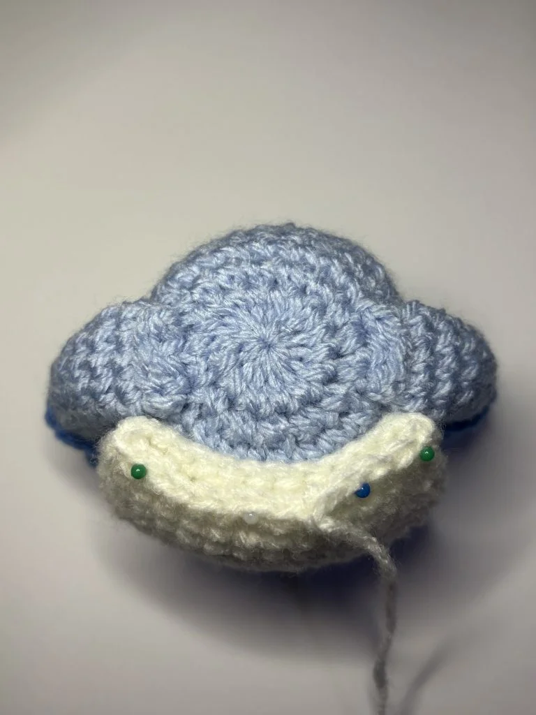 A crocheted blue and white object resembling a cloud or airplane, with pins and a loose yarn strand, placed on a plain white background—perfect inspiration for your next project, like a crochet gnome or free crochet pumpkin pattern.