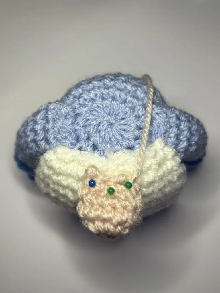 A crocheted blue and white turtle with a beige head, featuring three colorful pins and a loose yarn strand on its shell—a charming desk buddy for anyone who loves a good crochet gnome or unique crochet pumpkin pattern.