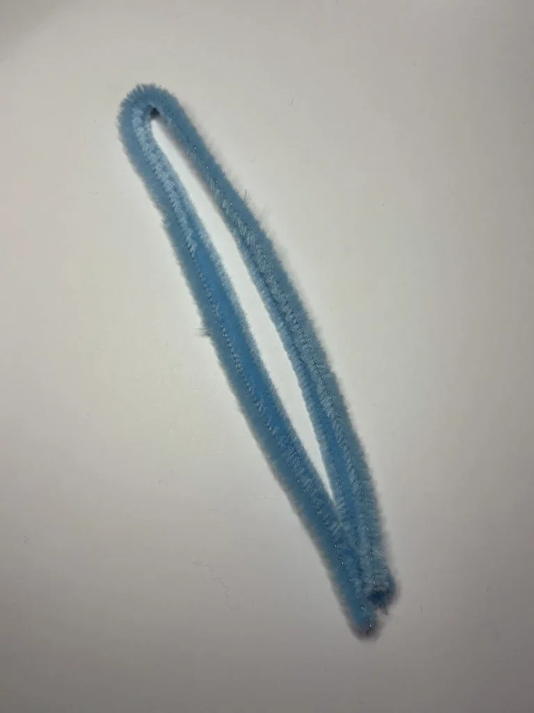 A single blue pipe cleaner bent into a narrow loop lies on a plain white surface, resembling the stem from a crochet pumpkin pattern.