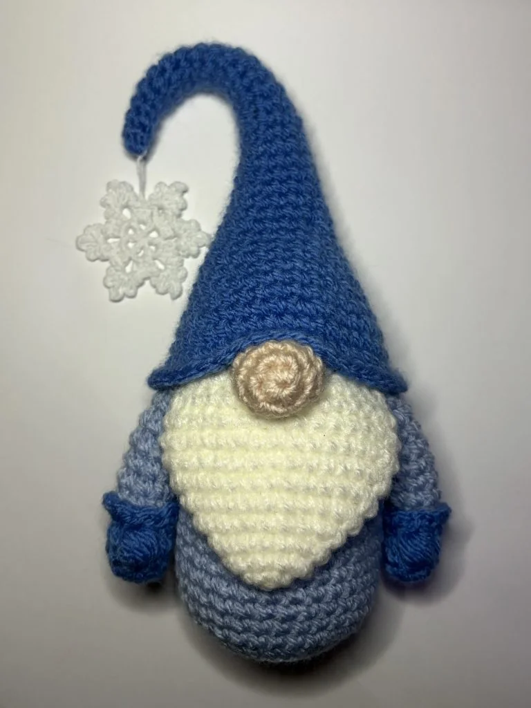 A charming crochet gnome doll with a long blue hat, white beard, blue mittens, and a crocheted white snowflake attached to the hat—perfect for those who love unique crochet gnome projects or are searching for a free crochet pumpkin pattern.