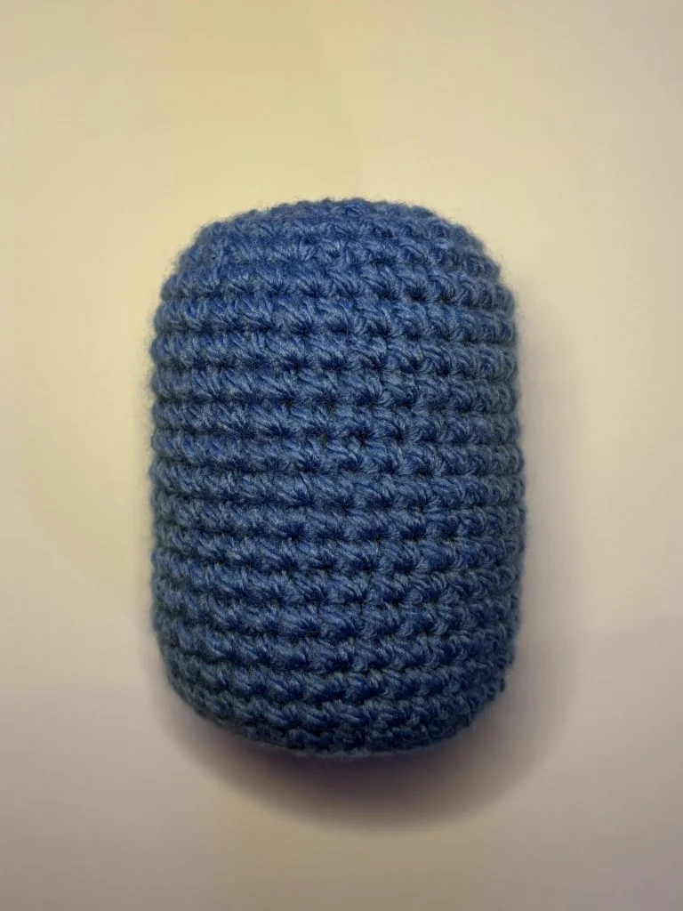 A small, blue, cylindrical crocheted object, reminiscent of a crochet gnome, is placed on a plain white surface.