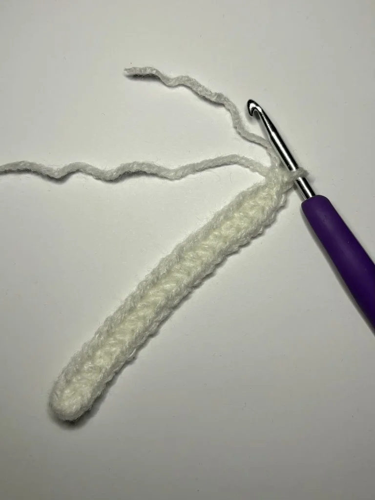 A partially crocheted white yarn piece, possibly the start of a pumpkin crochet pattern, with a purple-handled crochet hook attached, on a white background.