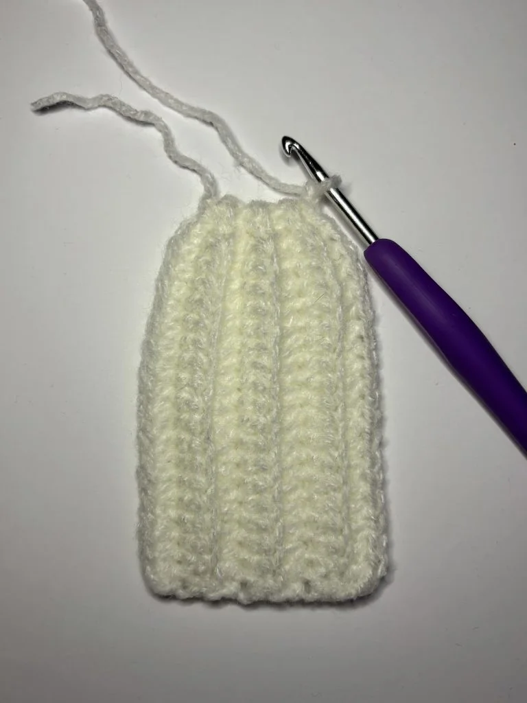 Crocheted white rectangular piece with vertical ridges, unfinished with a loose yarn end, and a metal crochet hook with a purple handle beside it—perfect for starting your next pumpkin crochet pattern.