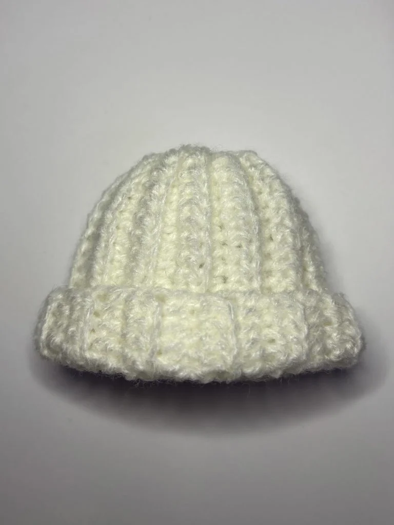 A small, white, knitted beanie hat with a folded brim, reminiscent of a cozy crochet gnome style, displayed on a plain white background.