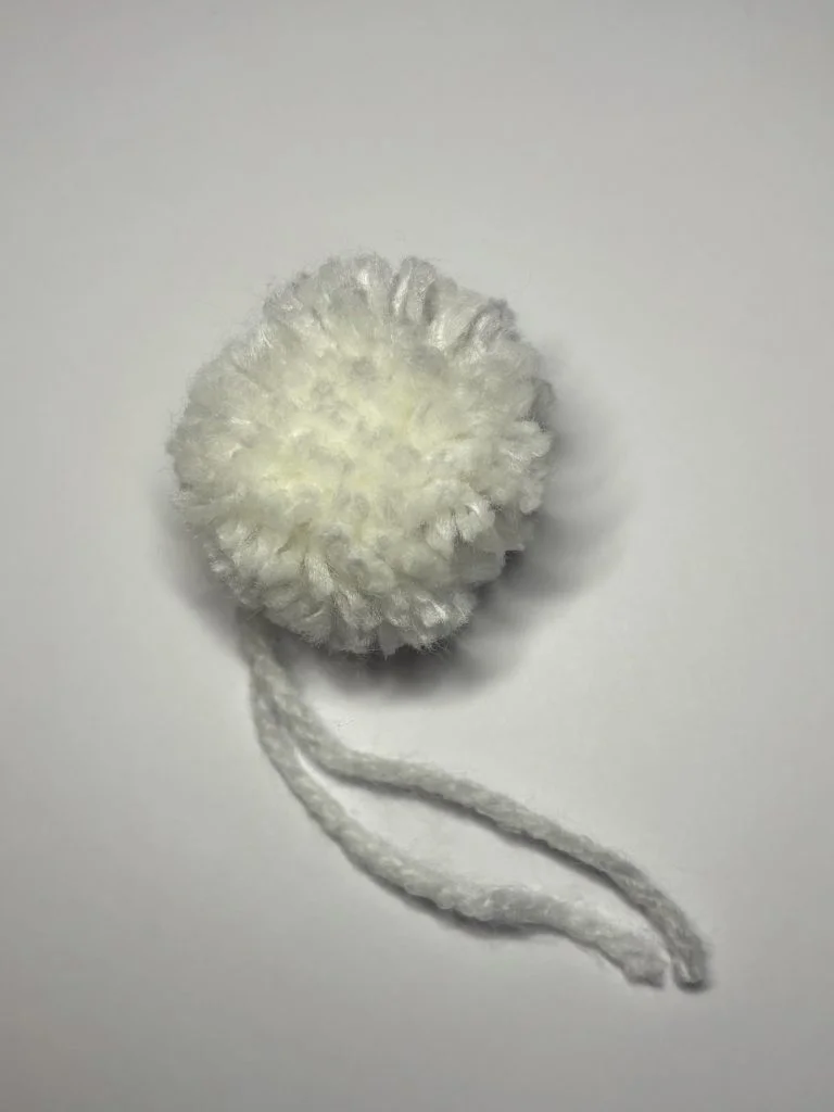 A small white yarn pom-pom with a loose string attached, placed on a plain white background, perfect for adding to your favorite pumpkin crochet pattern or other creative crochet projects.