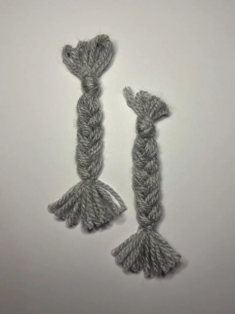 Two short braids made of thick gray yarn are arranged side by side on a plain white background, perfect for adding to a cute crochet gnome or as an accent for your pumpkin crochet pattern.