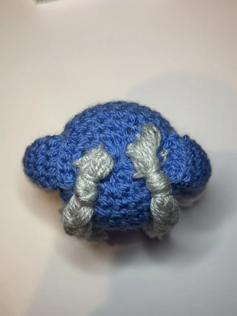 A small crocheted blue object made using a pumpkin crochet pattern, with two gray twisted strands attached on top, photographed on a white surface.