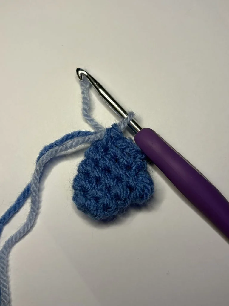 A metal crochet hook with a purple handle is attached to a partially crocheted blue yarn heart, resting on a white surface—perfect for practicing before trying a pumpkin crochet pattern.