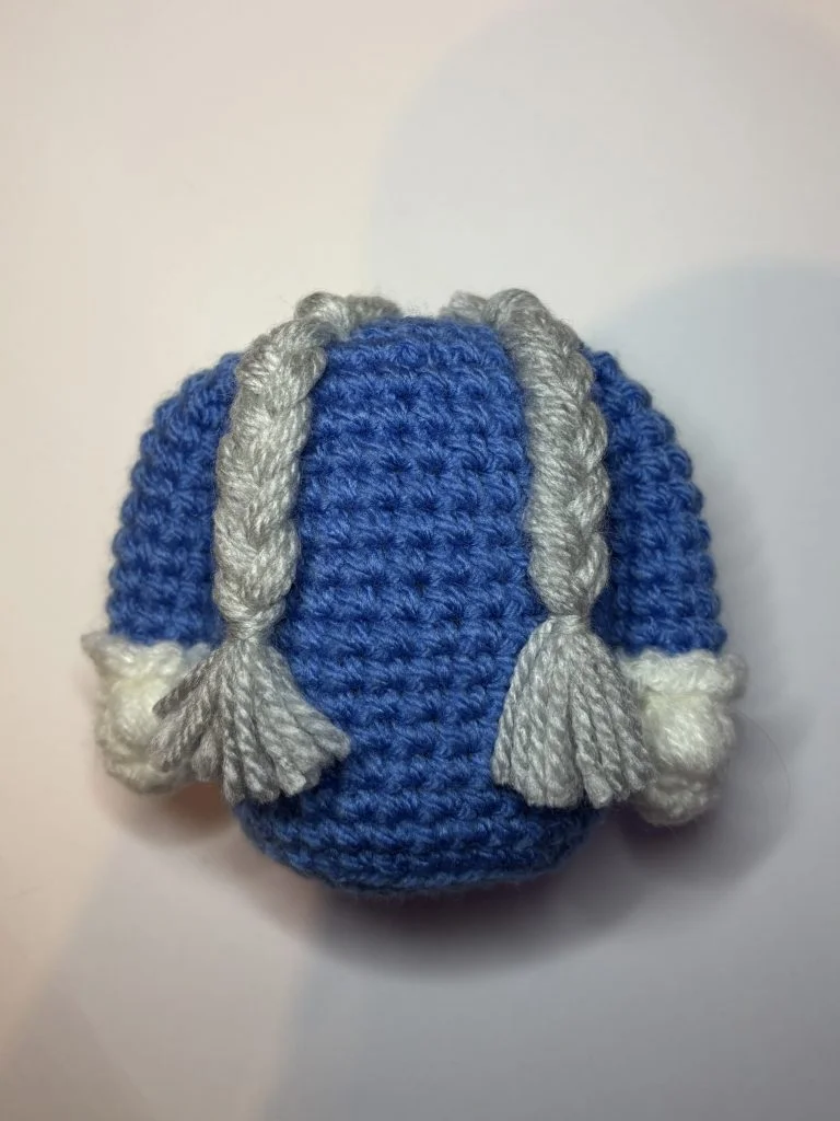A small crocheted blue sweater with white cuffs and two gray braided strands attached, perfect for pairing with your favorite free crochet pumpkin pattern.