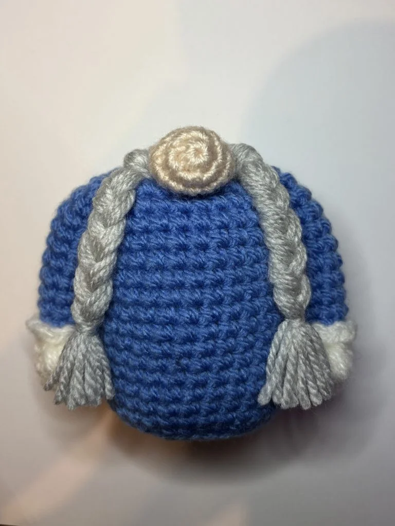 A crocheted blue object with long gray braids, white cuffs, and a beige circular section at the top, resembling a stylized character or figure—perfect inspiration if you're searching for a unique crochet pumpkin pattern.