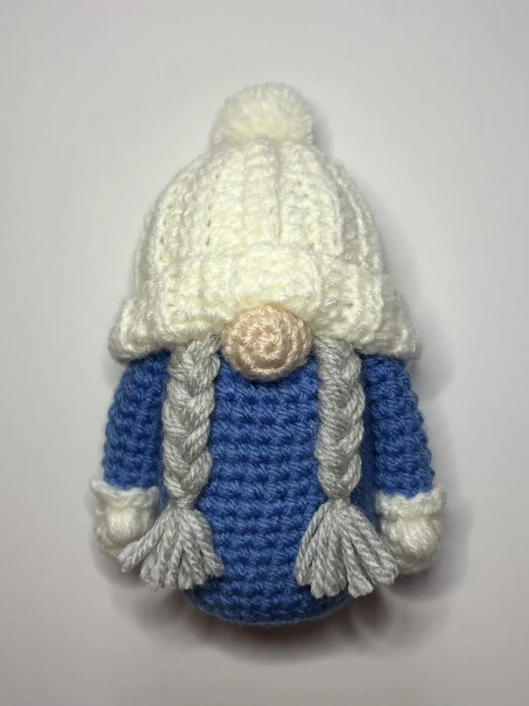 Crocheted doll with long gray braids, a blue body, white mittens, and a white hat with a pom-pom, resembling a charming crochet gnome. Photographed on a white background.
