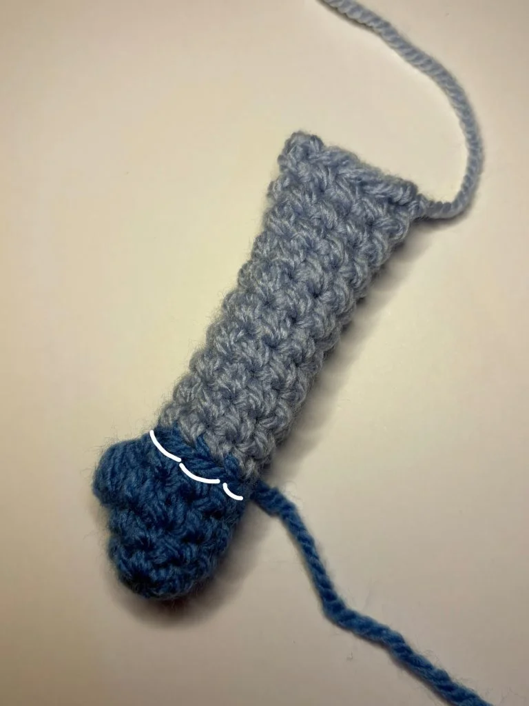 A crocheted object made with light and dark blue yarn, shaped like a small tube with a rounded end, reminiscent of a crochet gnome, lies on a plain surface.