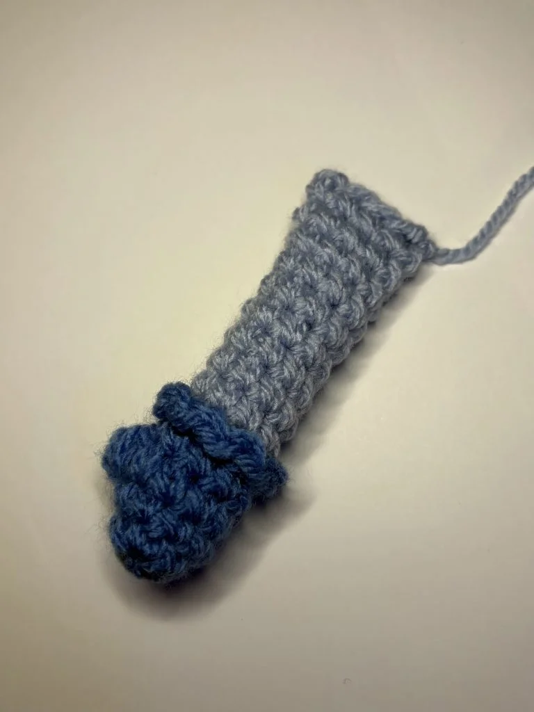 A small, crocheted object made with light blue and dark blue yarn, resembling a mitten or glove, lies on a plain white surface—perfect for practicing a pumpkin crochet pattern.