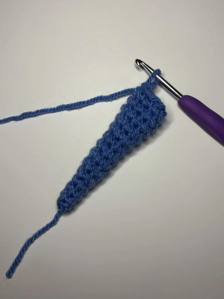 A blue crocheted cone-shaped piece with a loose yarn end and a crochet hook attached, placed on a plain white surface—perfect for starting your own free crochet pumpkin pattern.