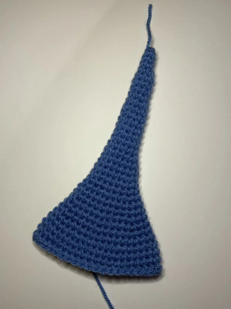A blue crocheted piece with a wide base that tapers to a narrow tip, reminiscent of a unique pumpkin crochet pattern, displayed on a plain white surface.