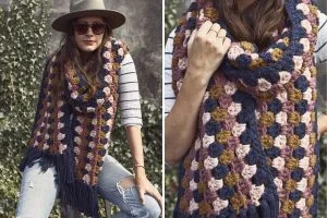 A woman wearing a wide-brim hat, sunglasses, and a striped shirt poses outdoors with a Super Scarf—a chunky, multi-colored crochet accessory featuring navy, pink, and mustard squares for a bold Color Pop.
