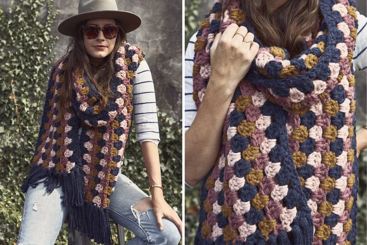 A woman wearing a wide-brim hat, sunglasses, and a striped shirt poses outdoors with a Super Scarf—a chunky, multi-colored crochet accessory featuring navy, pink, and mustard squares for a bold Color Pop.