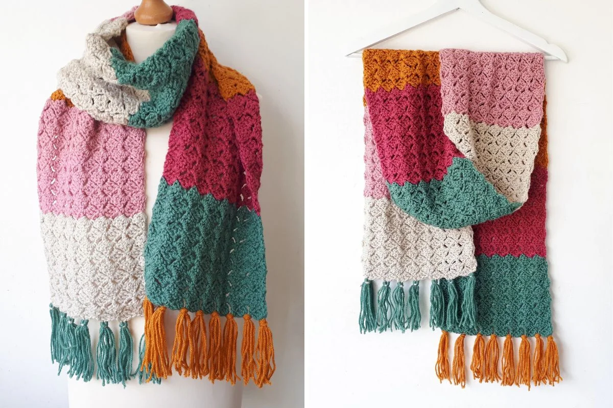 A multicolored crocheted scarf with pink, cream, teal, and orange sections, featuring textured patterns and orange fringe at the ends, shown draped and hanging.