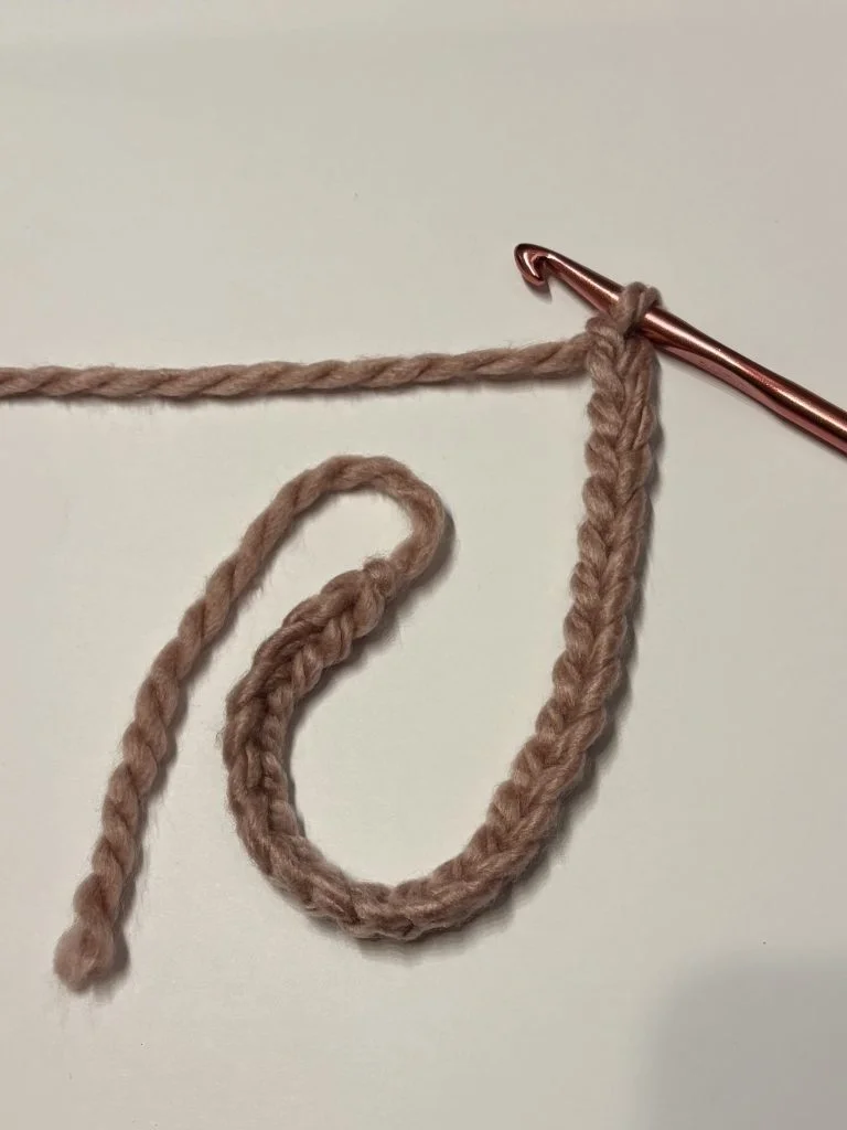 A copper crochet hook and pink yarn forming a foundation chain with several stitches on a white surface, perfect for starting a cozy crochet headband.