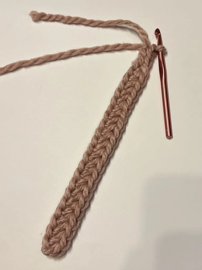 A pink crochet hook is attached to a short, narrow strip of pink crochet stitches on a white surface, showing the beginning of a crochet headband.