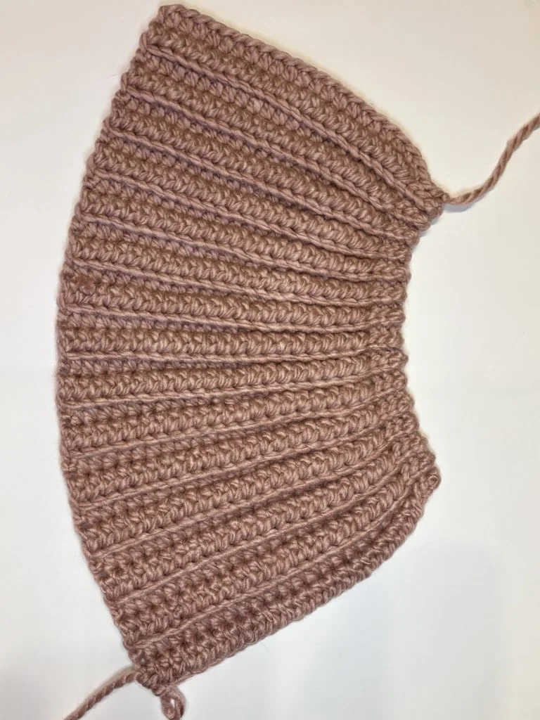 A pink ribbed crochet headband with curved edges lies flat on a white surface, showing yarn tails at both ends.