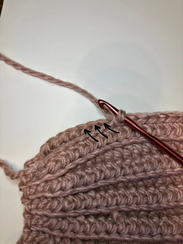 Close-up of a crochet headband project with pink yarn and hook, showing stitches and drawn arrows indicating where to insert the hook next.
