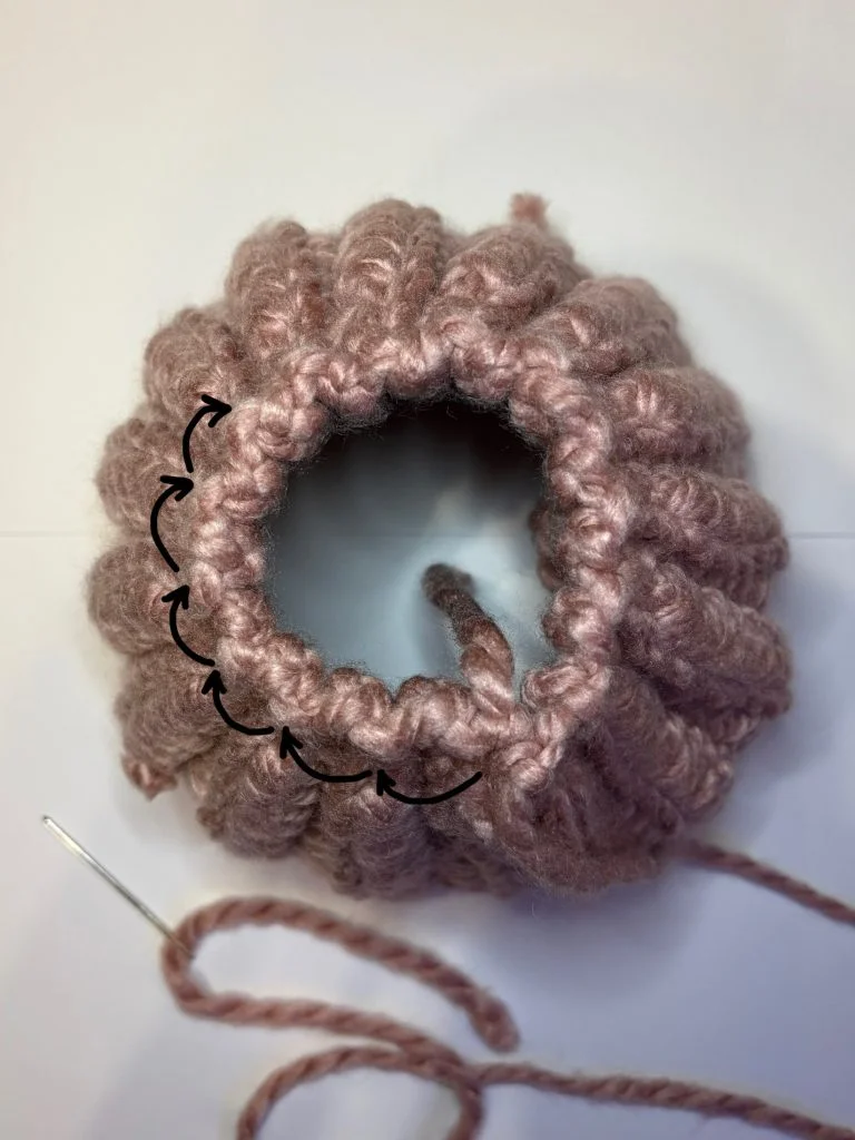 Close-up of a circular piece of pink crochet, possibly a crochet headband, with gathering yarn and needle, showing a partially closed opening and directional arrows indicating stitching.