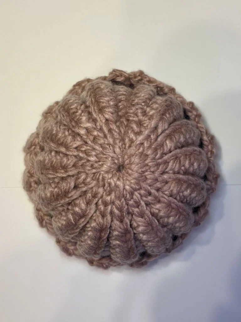 A round, pinkish-brown crochet headband with a ribbed pattern is viewed from above, displayed on a plain white surface.