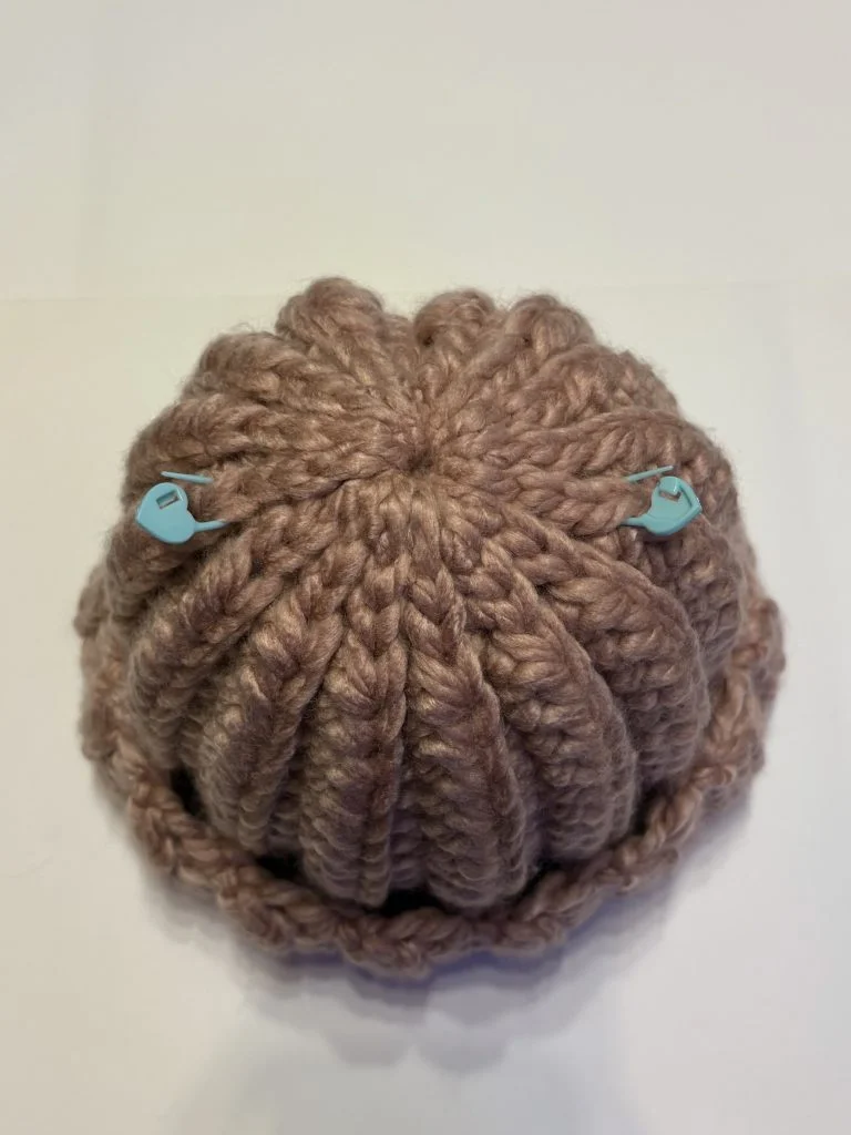 A thick, ribbed, brown crochet beanie lies flat, with two blue plastic stitch markers clipped to the top—perfect for pairing with your favorite crochet headband.